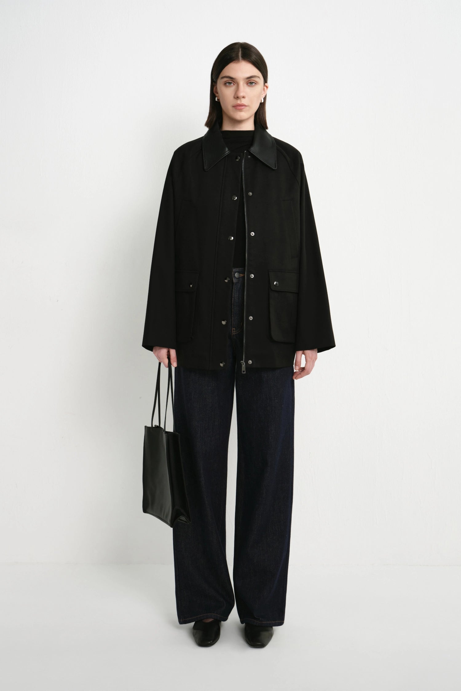 A model stands against a plain white studio background wearing a black button-front jacket over a black top, dark wide-leg pants, and black shoes, while holding a black tote bag.