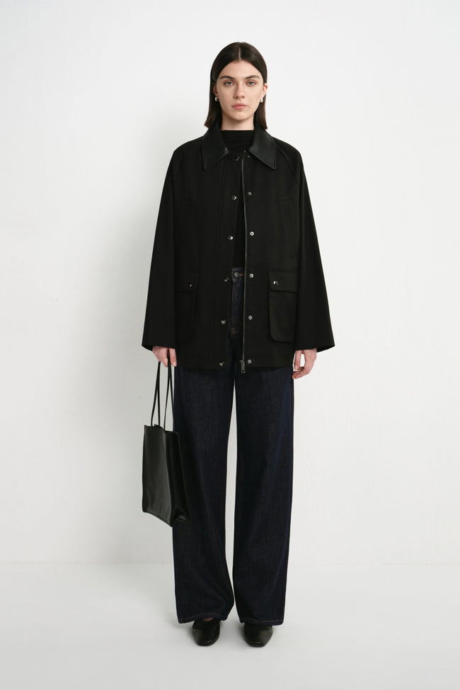 A model stands against a plain white studio background wearing a black button-front jacket over a black top, dark wide-leg pants, and black shoes, while holding a black tote bag.
