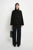 A model stands against a plain white studio background wearing a black button-front jacket over a black top, dark wide-leg pants, and black shoes, while holding a black tote bag.