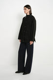 A model stands in profile wearing a black button-front jacket with a high collar, dark wide-leg trousers, and black shoes against a plain white studio background.