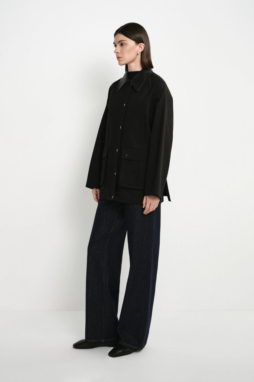 A model stands in profile wearing a black button-front jacket with a high collar, dark wide-leg trousers, and black shoes against a plain white studio background.