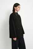 A model shown in profile wearing a black button-front jacket with a structured collar and dark wide-leg pants, photographed against a plain light gray studio background.