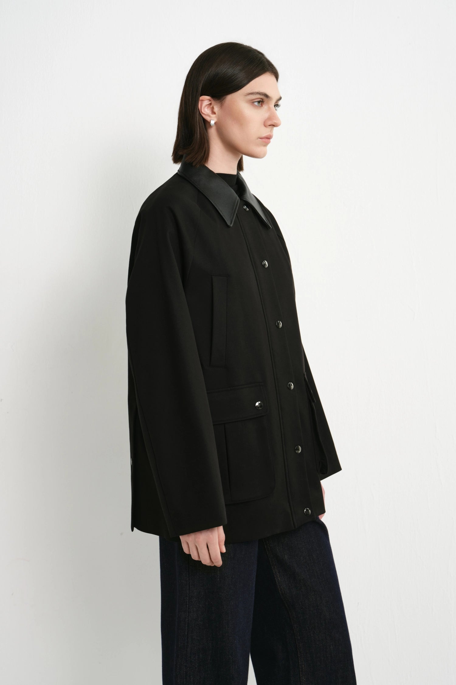 A model shown in profile wearing a black button-front jacket with a structured collar and dark wide-leg pants, photographed against a plain light gray studio background.