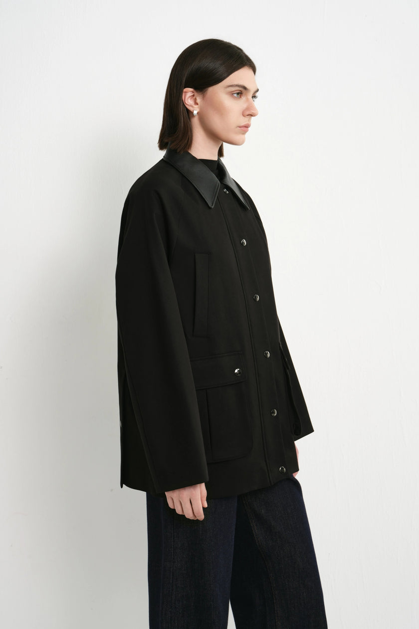 A model shown in profile wearing a black button-front jacket with a structured collar and dark wide-leg pants, photographed against a plain light gray studio background.