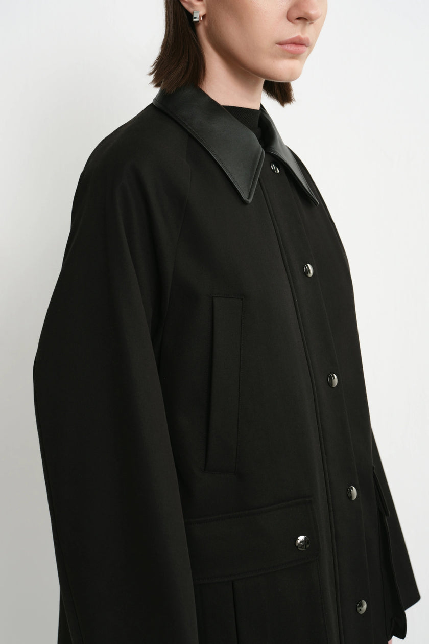 A close-up side view of a model wearing a black oversized button-front jacket with a contrasting black collar, shown against a plain light gray studio background.