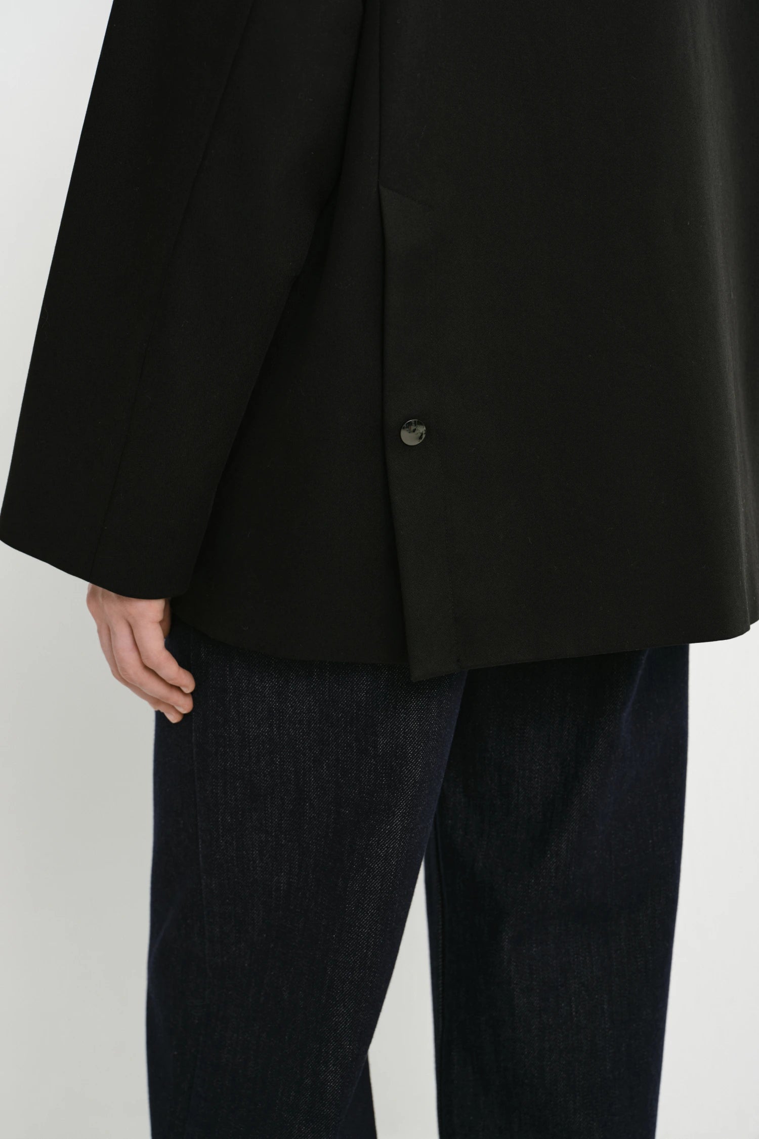 Close-up of a person wearing a black oversized blazer with a single visible button and dark blue jeans against a light gray studio background.