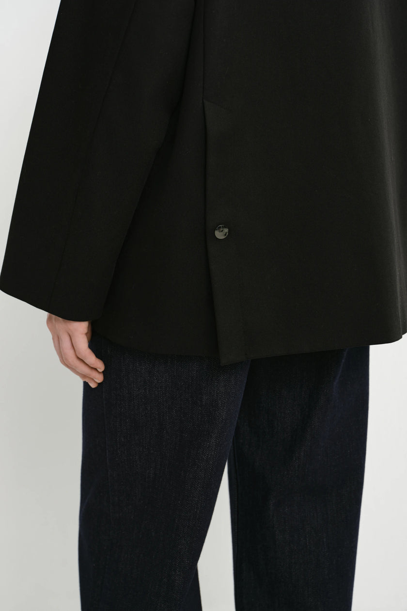 Close-up of a person wearing a black oversized blazer with a single visible button and dark blue jeans against a light gray studio background.