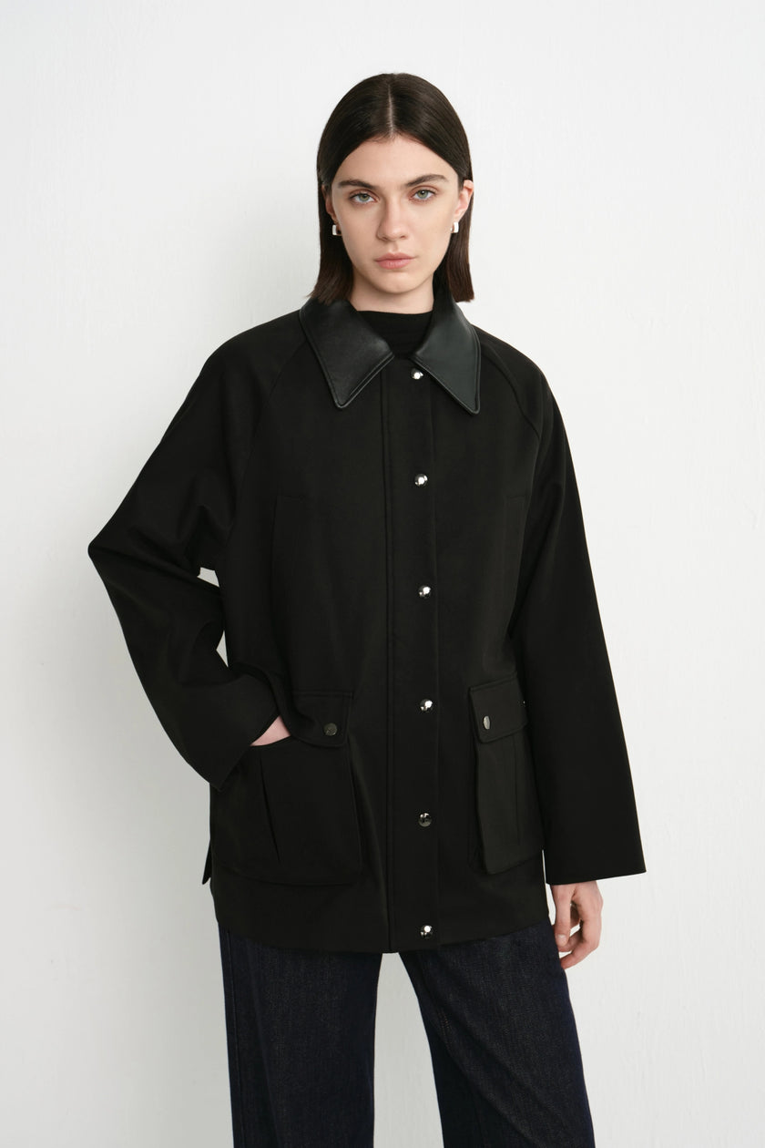 A model wearing a black utility jacket with contrasting black leather-like collar and button details, paired with dark jeans, against a plain light gray studio background.