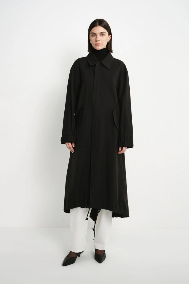 A model stands against a plain white studio background wearing a long black coat over a black turtleneck, white wide-leg pants, and black pointed shoes.