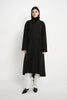 A model stands against a plain white studio background wearing a long black coat over a black turtleneck, white wide-leg pants, and black pointed shoes.