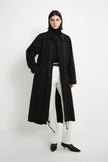 A model stands against a plain white studio background wearing a long black trench coat over a black turtleneck, white wide-leg trousers, and black pointed-toe heels.