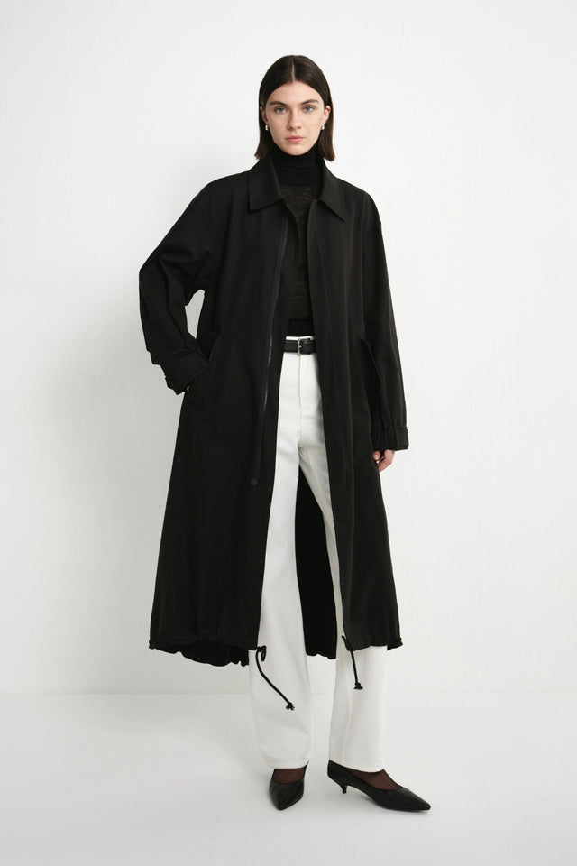 A model stands against a plain white studio background wearing a long black trench coat over a black turtleneck, white wide-leg trousers, and black pointed-toe heels.