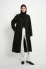 A model stands against a plain white studio background wearing a long black trench coat over a black turtleneck, white wide-leg trousers, and black pointed-toe heels.