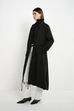 A model stands in profile wearing a long black coat over a white turtleneck top and white pants, paired with black pointed shoes, against a plain white studio background.