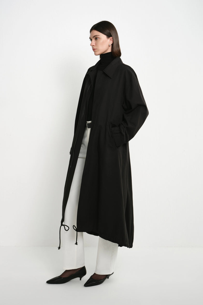 A model stands in profile wearing a long black coat over a white turtleneck top and white pants, paired with black pointed shoes, against a plain white studio background.