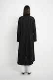 A model viewed from the back wearing a long black coat over white pants and black shoes, standing against a plain light gray studio background.