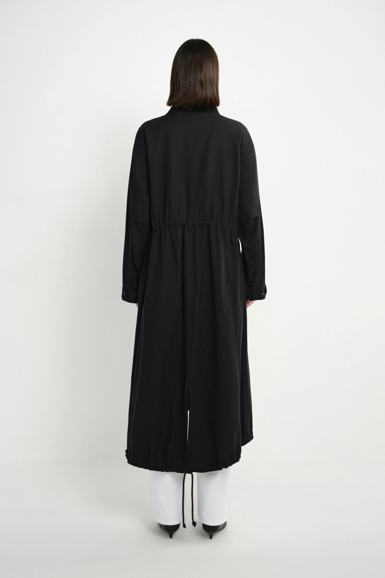 A model viewed from the back wearing a long black coat over white pants and black shoes, standing against a plain light gray studio background.