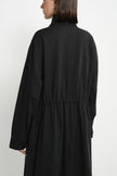 A woman shown from the back wearing a long black coat or dress with a gathered waist seam, photographed against a plain light background.