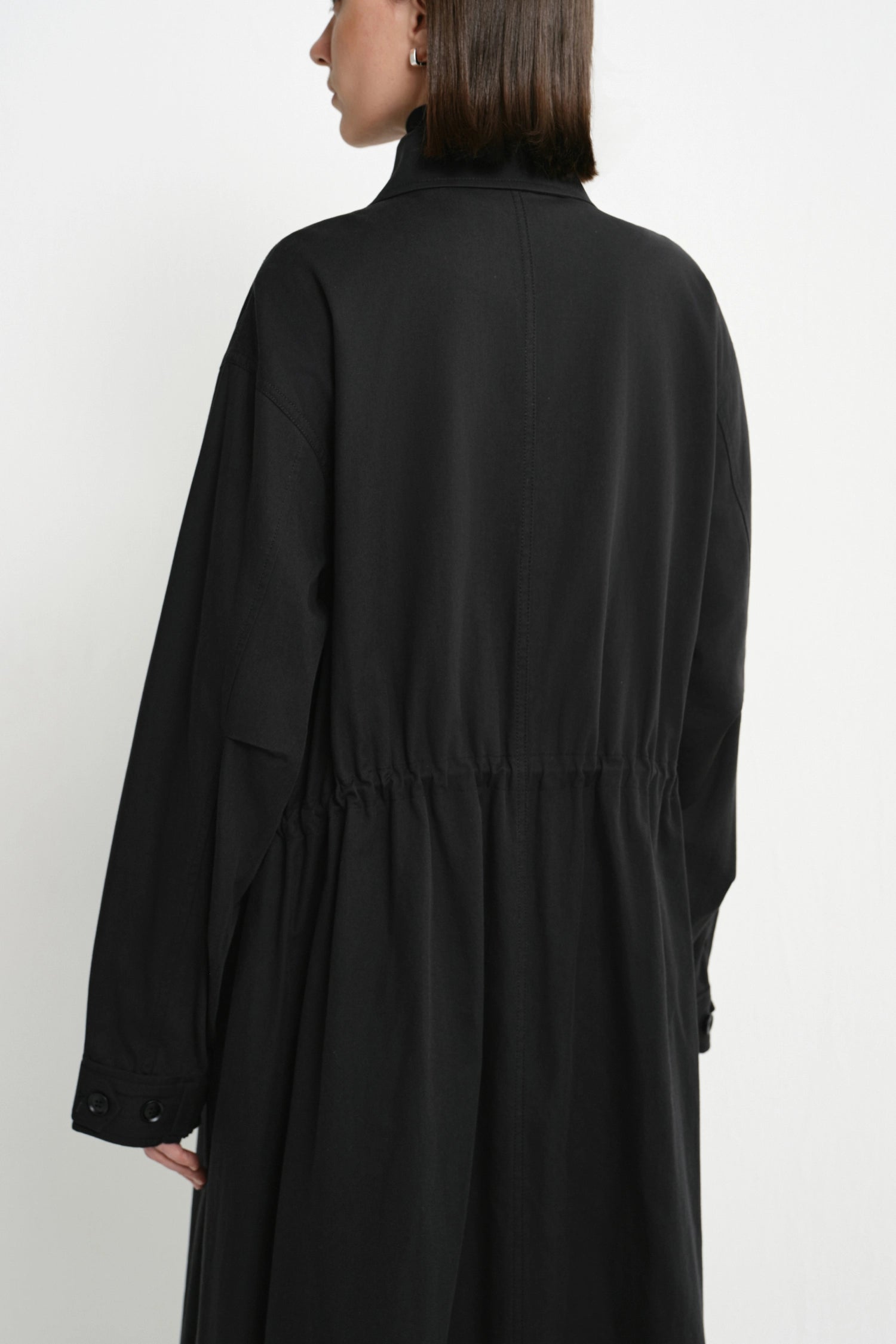 A woman shown from the back wearing a long black coat or dress with a gathered waist seam, photographed against a plain light background.