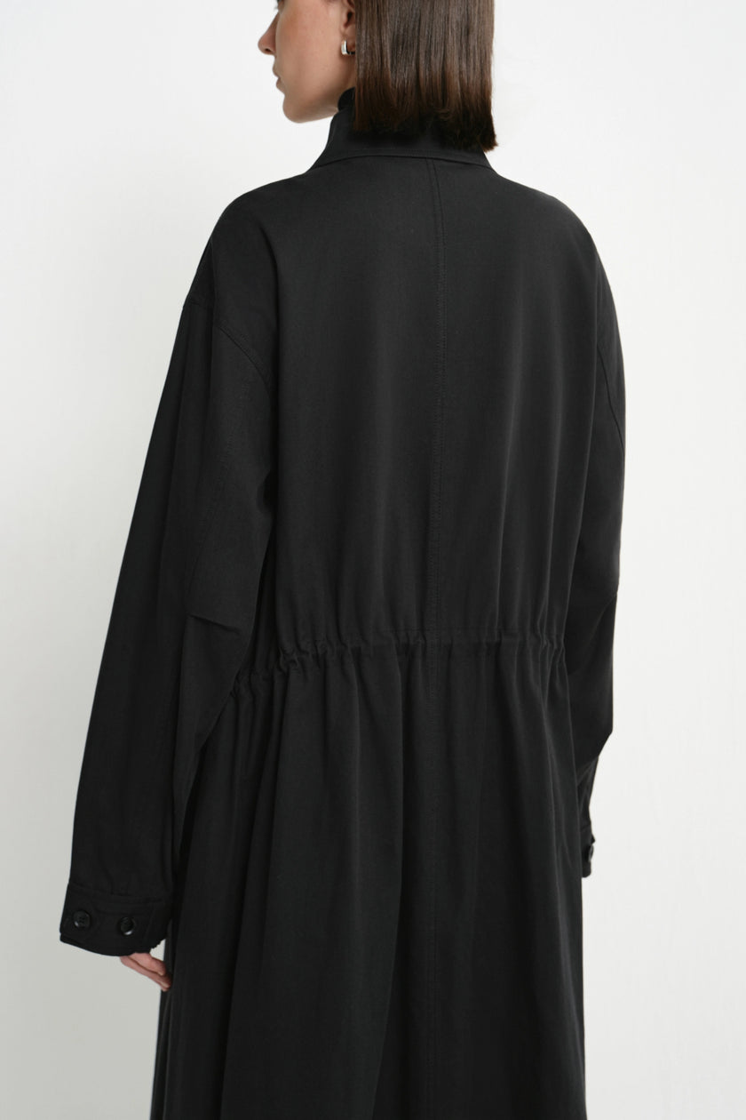 A woman shown from the back wearing a long black coat or dress with a gathered waist seam, photographed against a plain light background.
