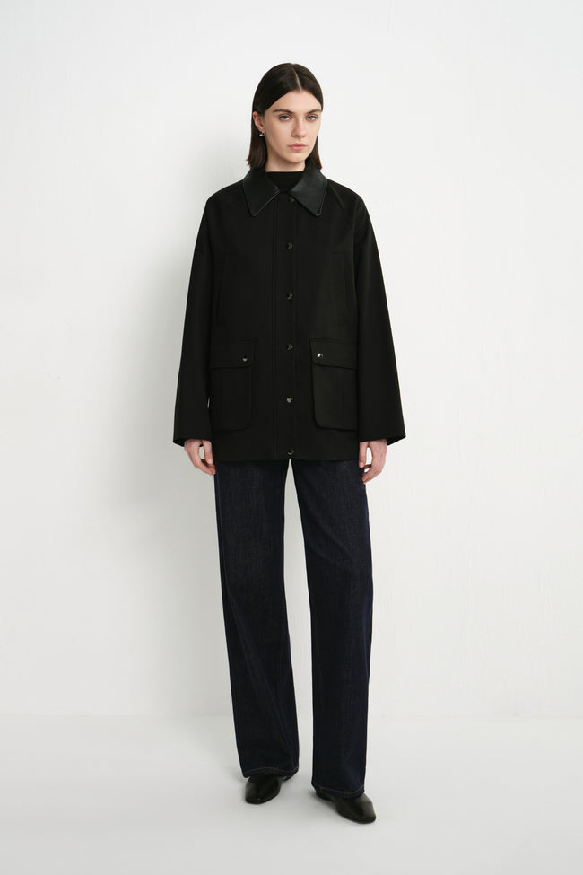 A model wearing the Rue Sophie Roth Leather Barn Jacket in black, featuring a contrast leather collar, metallic snap-front closure, utility pockets, and a relaxed hip-length silhouette, front view.