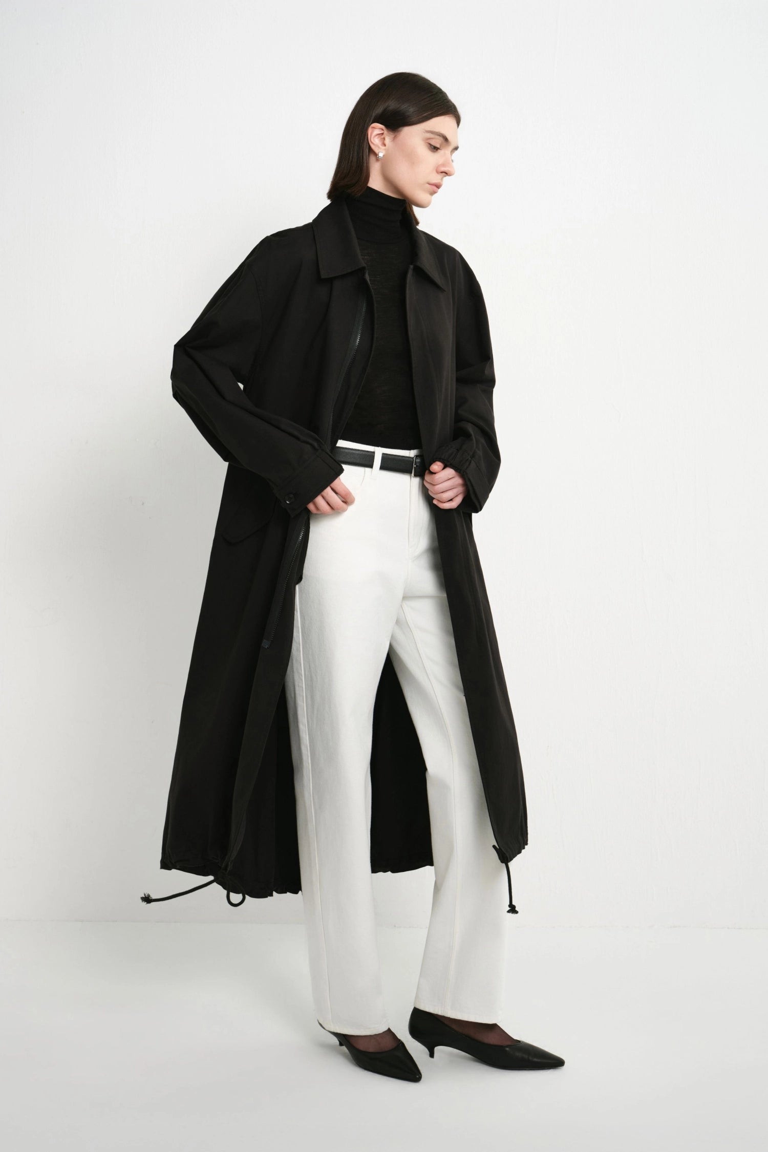 A model wearing a long black trench coat over a black turtleneck, white wide-leg trousers, and black pointed shoes, standing against a plain white studio background.