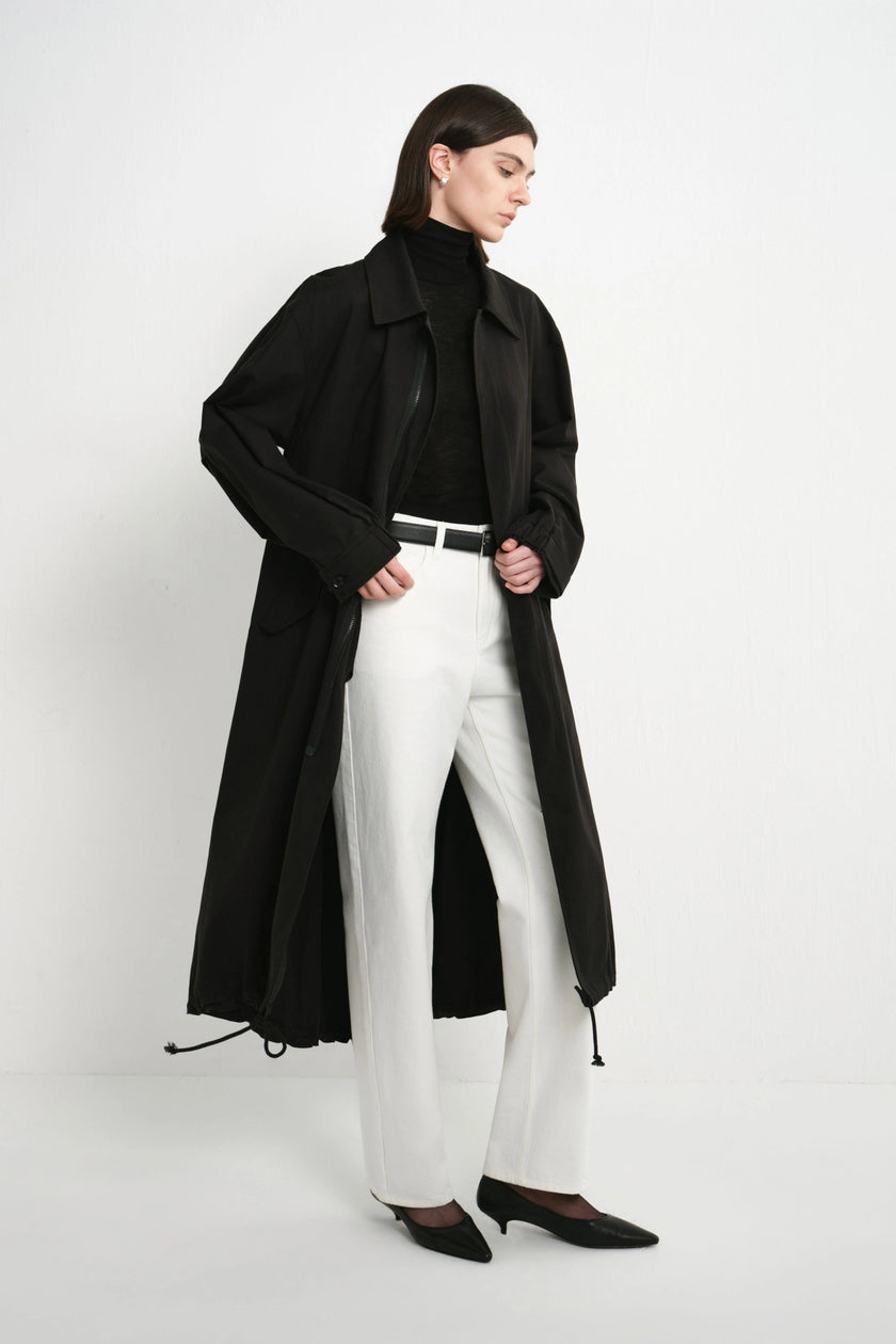 A model wearing a long black trench coat over a black turtleneck, white wide-leg trousers, and black pointed shoes, standing against a plain white studio background.
