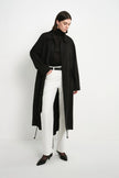A model wearing a long black trench coat over a black turtleneck, white trousers, and black pointed shoes, standing against a plain white studio background.