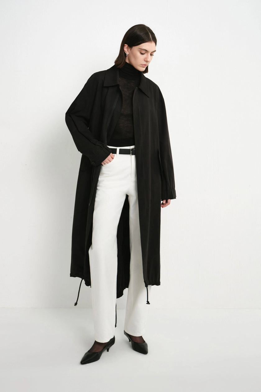 A model wearing a long black trench coat over a black turtleneck, white trousers, and black pointed shoes, standing against a plain white studio background.