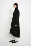 A model is shown in profile wearing a long black coat over white pants and black pointed shoes, standing against a plain white studio background.