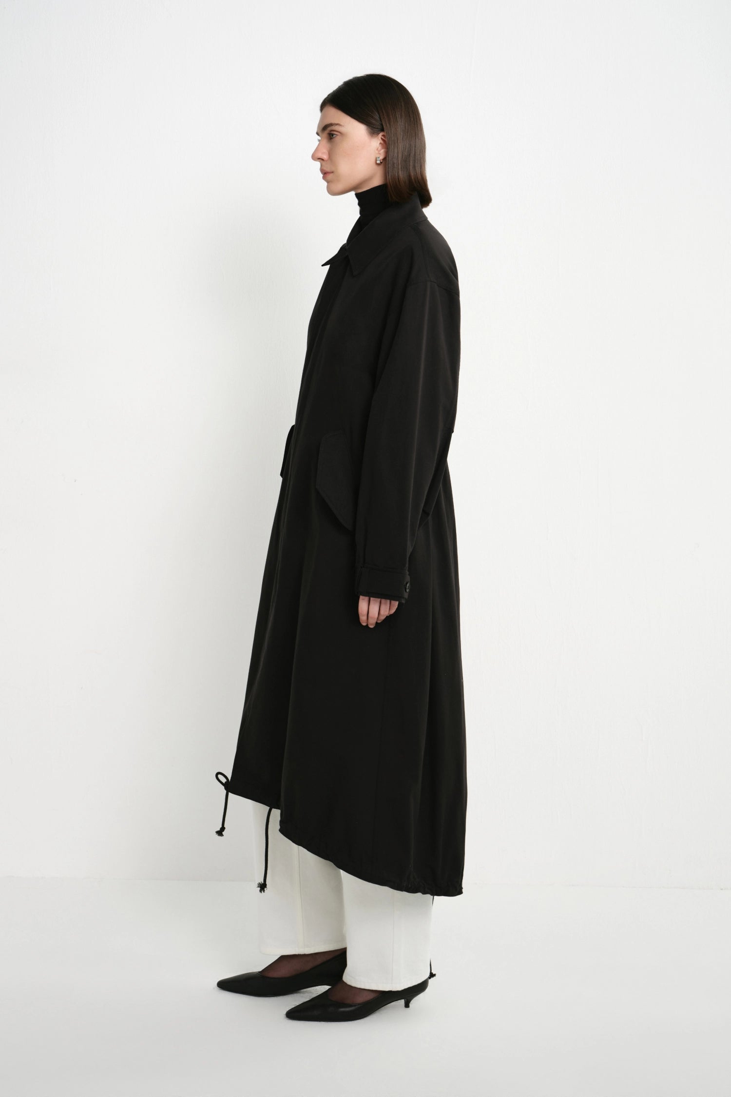 A model is shown in profile wearing a long black coat over white pants and black pointed shoes, standing against a plain white studio background.