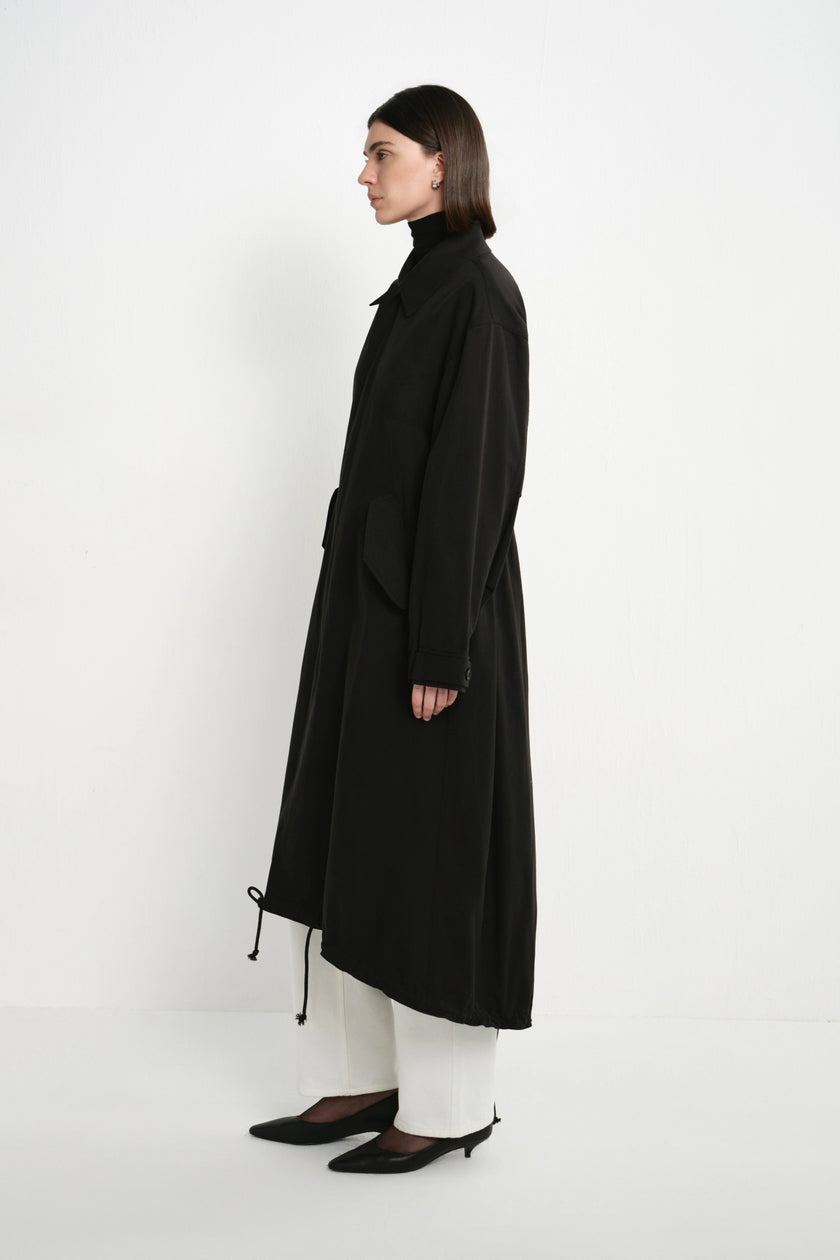 A model is shown in profile wearing a long black coat over white pants and black pointed shoes, standing against a plain white studio background.
