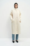 A model wearing the Rue Sophie Anne Satin Coat in crème with a high collar, metallic snap buttons, long raglan sleeves, and a clean straight silhouette, front view.
