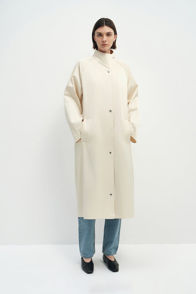 A model wearing the Rue Sophie Anne Satin Coat in crème with a high collar, metallic snap buttons, long raglan sleeves, and a clean straight silhouette, front view.