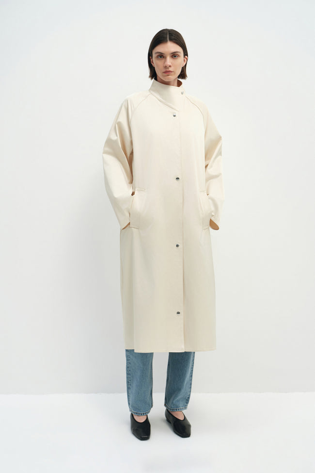 A model wearing the Rue Sophie Anne Satin Coat in crème with a high collar, metallic snap buttons, long raglan sleeves, and a clean straight silhouette, front view.