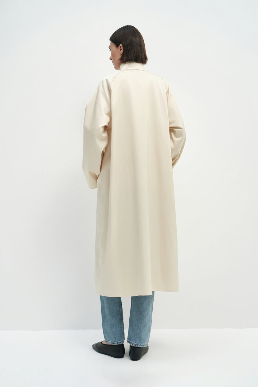 Back view of a model wearing the Rue Sophie Anne Satin Coat in crème, highlighting the high collar, raglan shoulder construction, long sleeves, and clean longline silhouette.