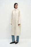 A model wearing the Rue Sophie Anne Satin Coat in crème, featuring a high stand collar, metallic snap closure, side pockets, and a relaxed long silhouette, three-quarter front view.