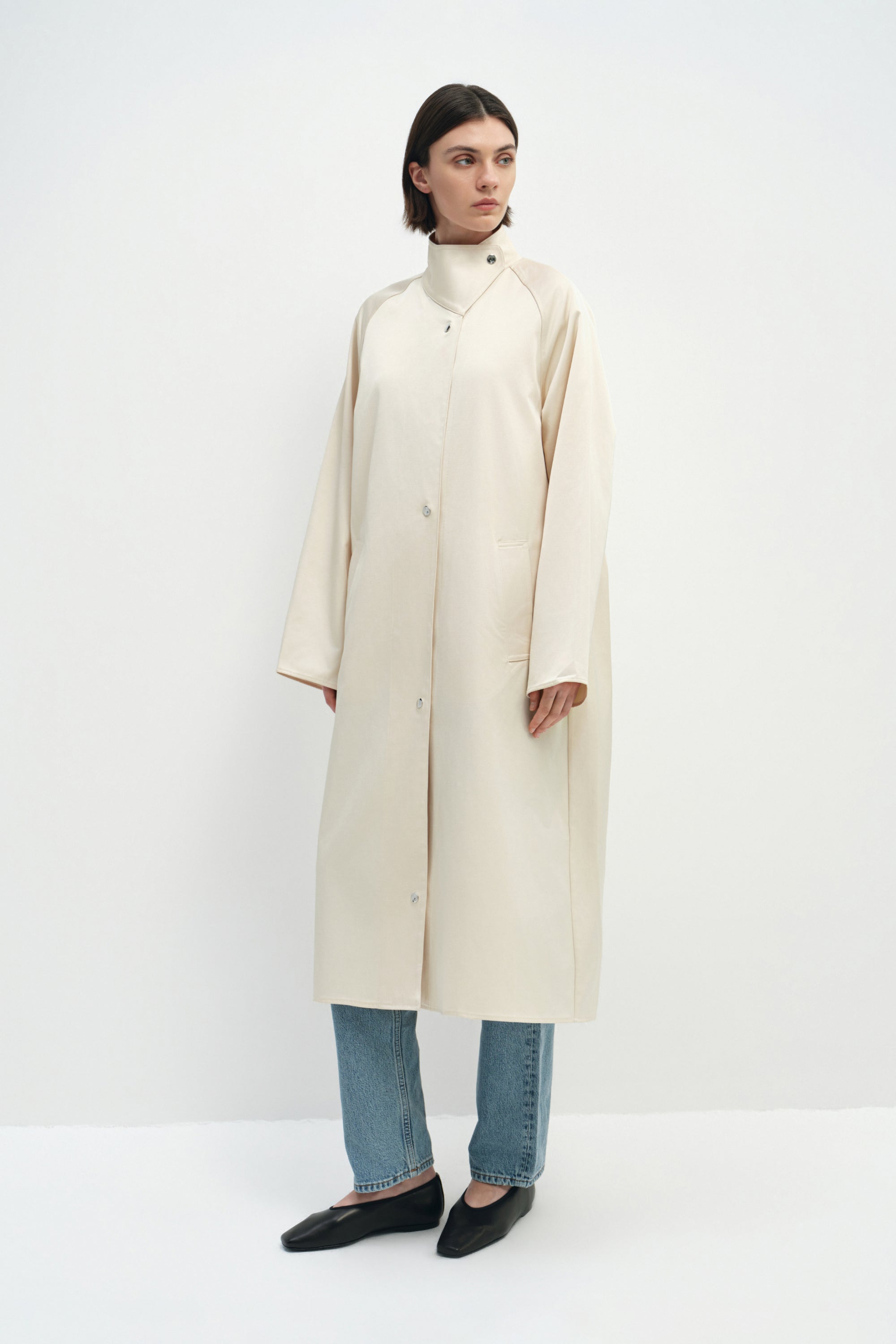 A model wearing the Rue Sophie Anne Satin Coat in crème, featuring a high stand collar, metallic snap closure, side pockets, and a relaxed long silhouette, three-quarter front view.