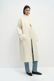 A model wearing the Rue Sophie Anne Satin Coat in crème with a high collar, metallic snap-front detailing, raglan sleeves, and a fluid longline shape, three-quarter view.