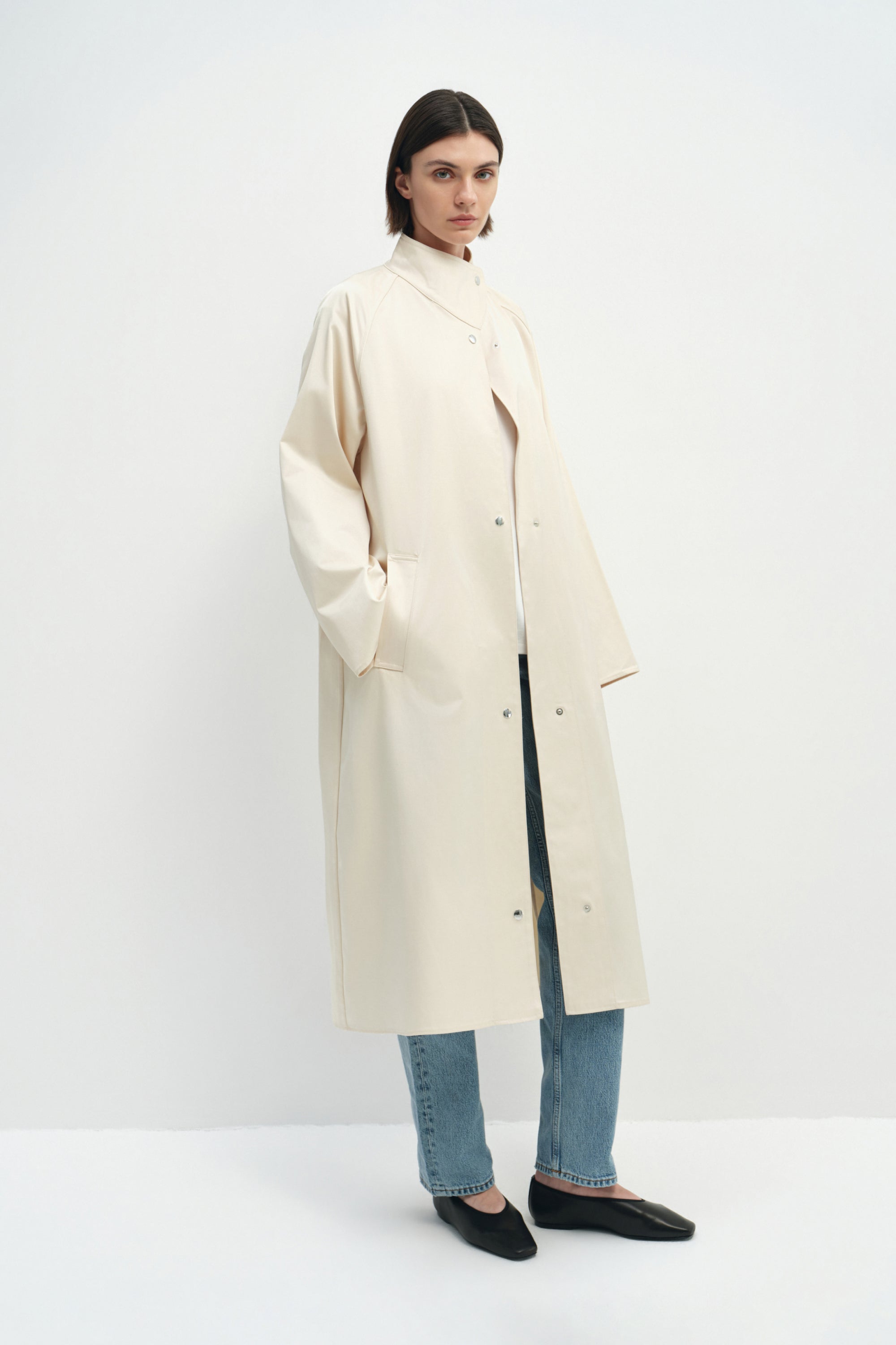 A model wearing the Rue Sophie Anne Satin Coat in crème with a high collar, metallic snap-front detailing, raglan sleeves, and a fluid longline shape, three-quarter view.