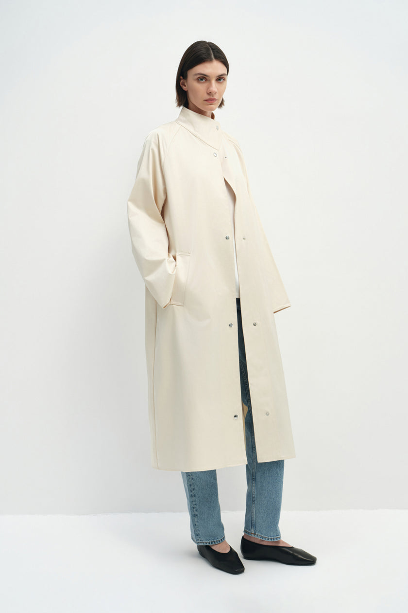 A model wearing the Rue Sophie Anne Satin Coat in crème with a high collar, metallic snap-front detailing, raglan sleeves, and a fluid longline shape, three-quarter view.