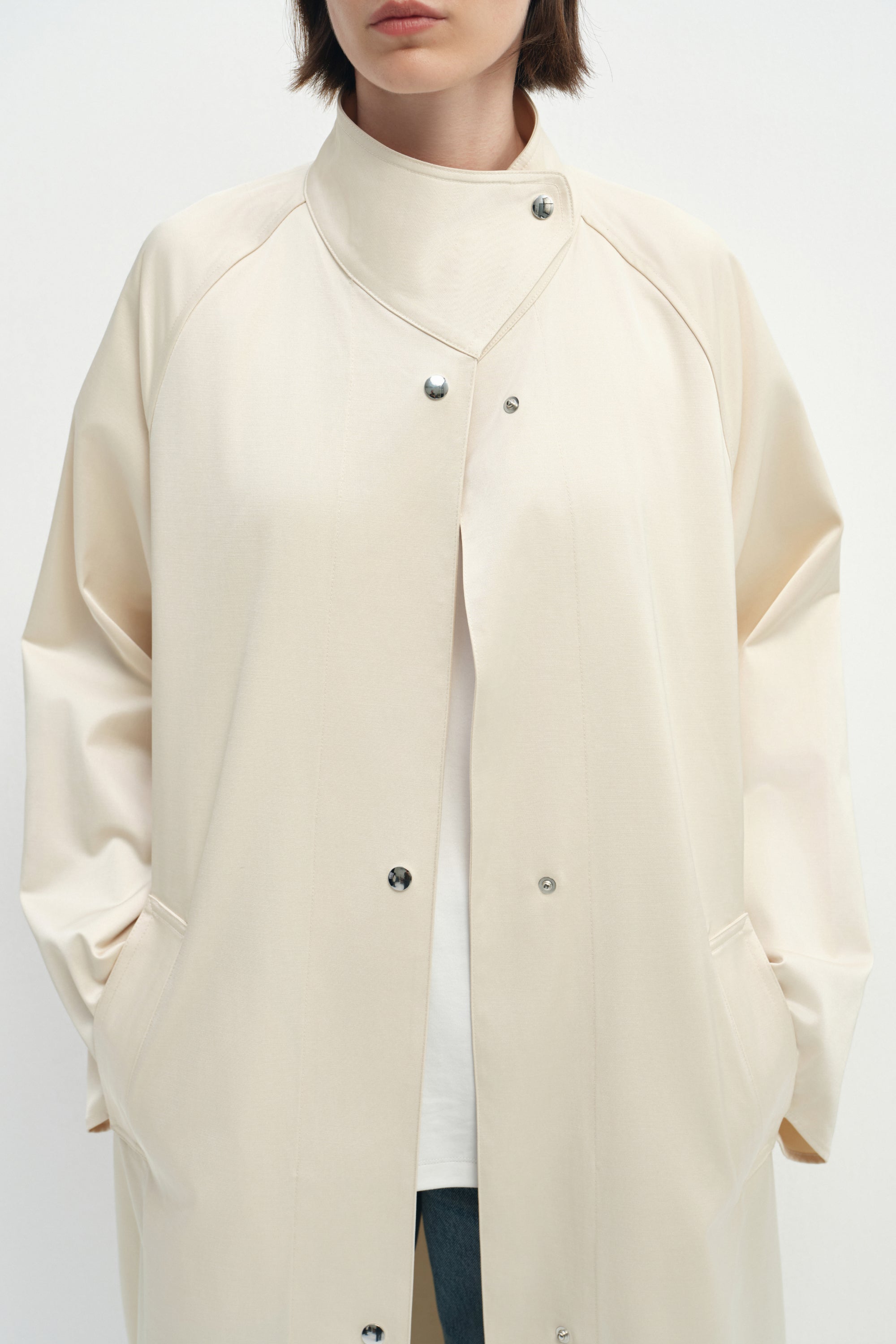 Close-up of the Rue Sophie Anne Satin Coat in crème, highlighting the high stand collar, metallic snap closures, raglan seam construction, side pockets, and smooth satin finish.