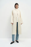 A model wearing the Rue Sophie Anne Satin Coat in crème, featuring a high stand collar, metallic snap-front closure, raglan sleeves, side pockets, and a relaxed longline silhouette, front view.