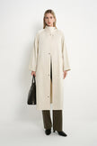 A model stands against a plain white studio background wearing a long cream-colored coat over a beige top, olive green wide-leg trousers, and black shoes, while holding a black leather handbag.