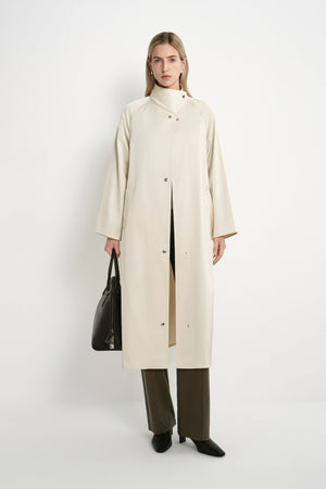 A model stands against a plain white studio background wearing a long cream-colored coat over a beige top, olive green wide-leg trousers, and black shoes, while holding a black leather handbag.