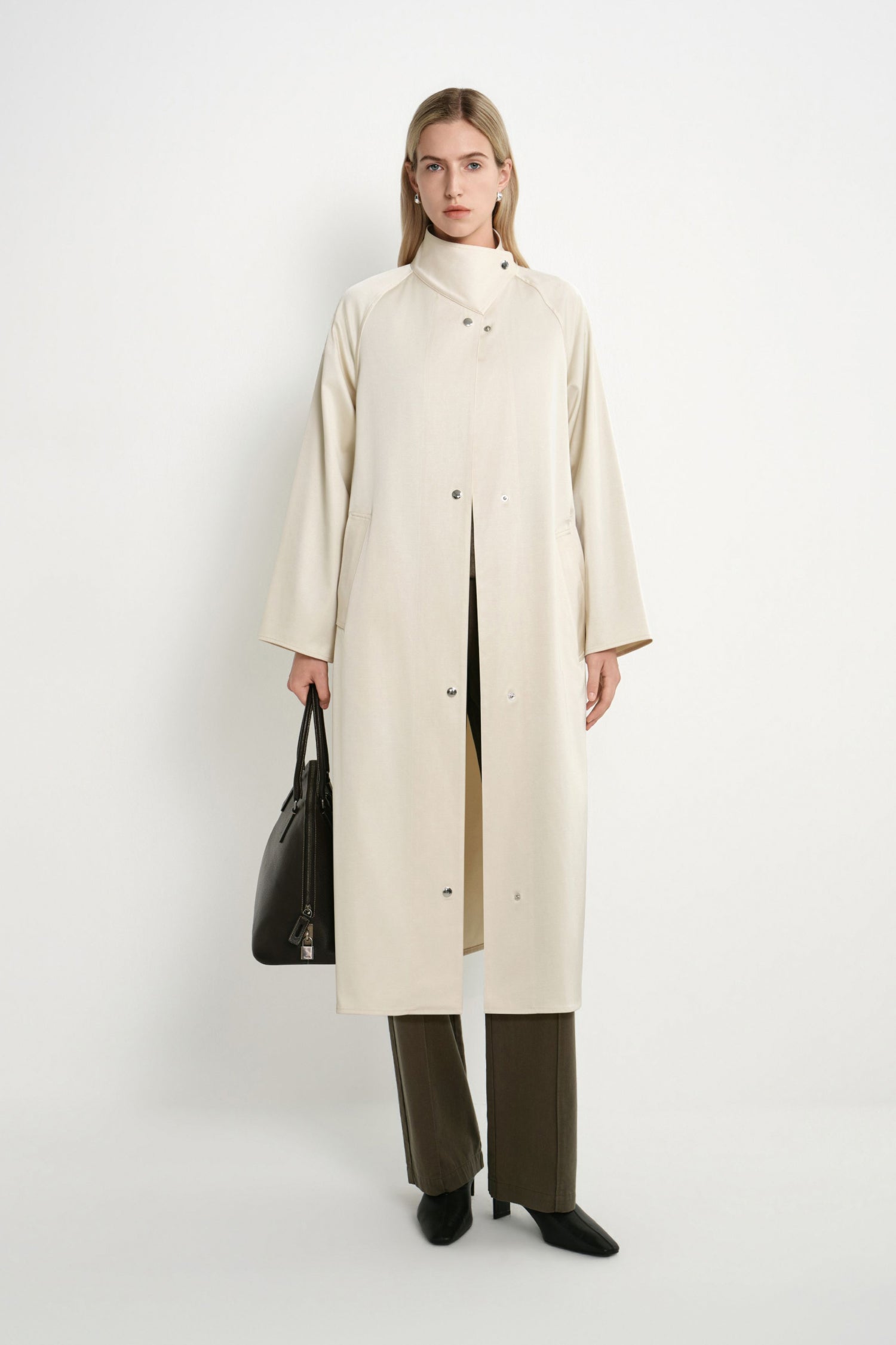 A model stands against a plain white studio background wearing a long cream-colored coat over a beige top, olive green wide-leg trousers, and black shoes, while holding a black leather handbag.