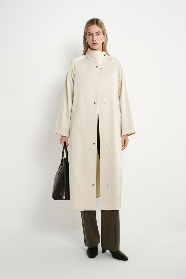 A model stands against a plain white studio background wearing a long cream-colored coat over a beige top, olive green wide-leg trousers, and black shoes, while holding a black leather handbag.