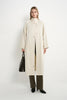 A model stands against a plain white studio background wearing a long cream-colored coat over a beige top, olive green wide-leg trousers, and black shoes, while holding a black leather handbag.