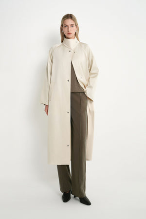 A model stands against a plain white studio background wearing a long cream-colored coat over a brown top and wide-leg brown trousers, paired with black pointed shoes.