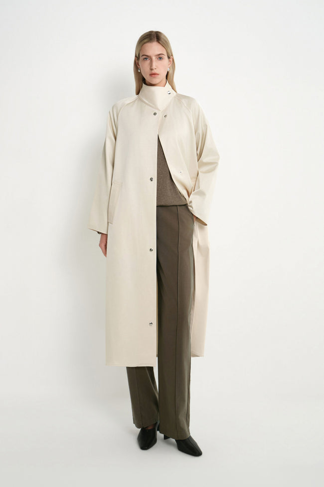A model stands against a plain white studio background wearing a long cream-colored coat over a brown top and wide-leg brown trousers, paired with black pointed shoes.